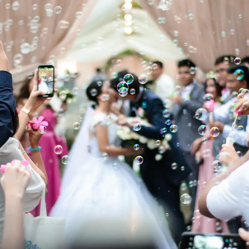 Hochzeitspaar küsst sich unter einem geschmückten Pavillon, während Gäste Seifenblasen pusten und den Moment mit Smartphones festhalten