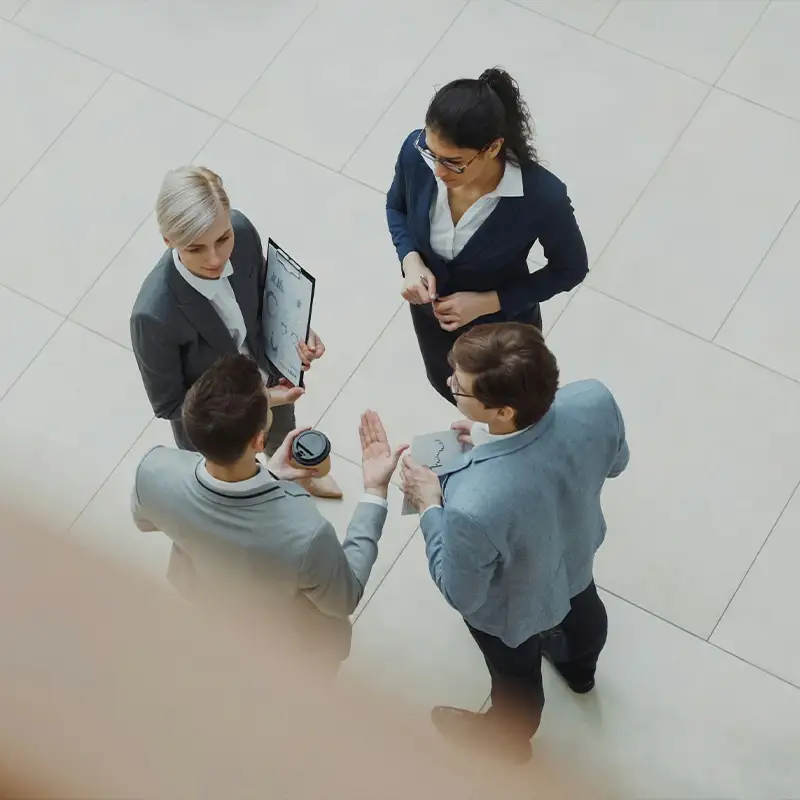 Vier Geschäftsleute stehen in einem offenen, hellen Raum im Gespräch beisammen, von oben fotografiert, eine Person hält ein Klemmbrett mit Diagramm