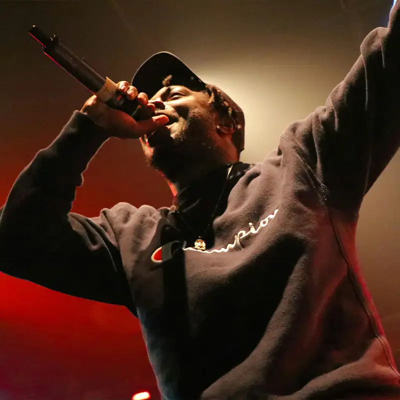 Rapper mit Mikrofon auf einer Bühne, trägt Sweatshirt und Basecap, dramatische Beleuchtung in Rot und Gelb von unten