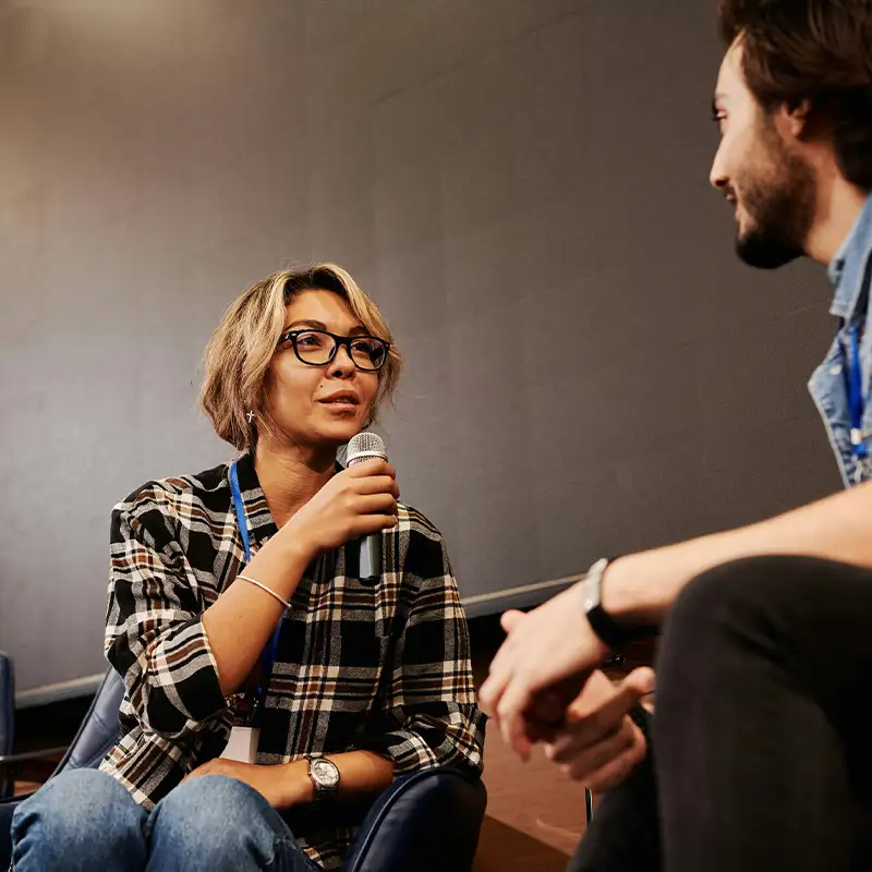 Frau mit Mikrofon im Gespräch mit einem Mann, beide sitzen in Sesseln und tragen Konferenz-Badges