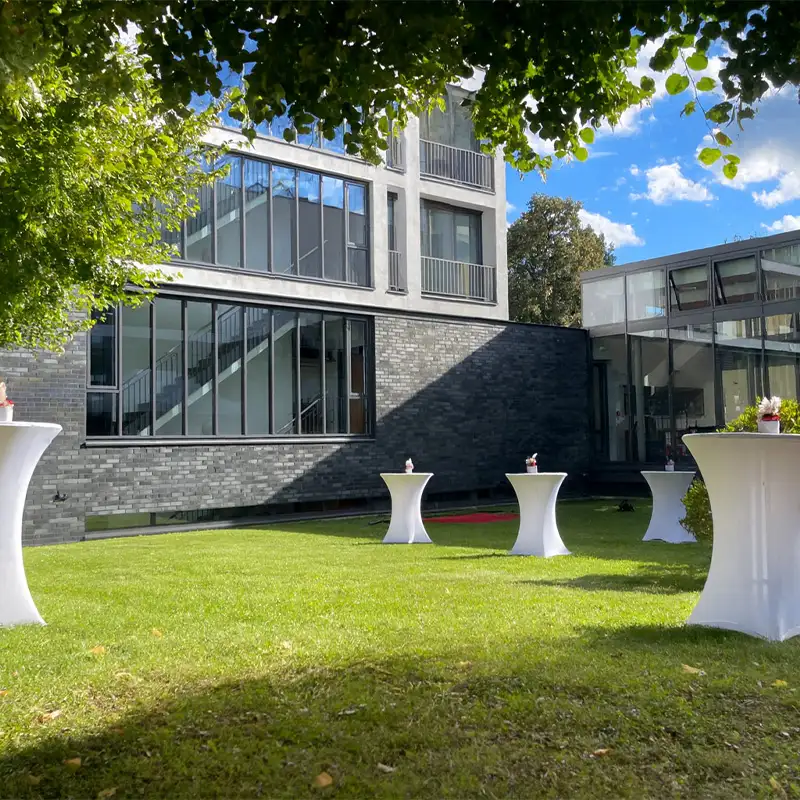 Moderner Gebäudekomplex mit Glas- und Ziegelfassade, davor eine gepflegte Rasenfläche mit mehreren weiß eingedeckten Stehtischen unter Bäumen.