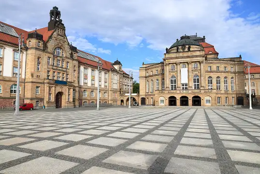 Weitläufiger Platz mit geometrischem Pflastermuster vor zwei prächtigen historischen Gebäuden mit Sandsteinfassaden und roten Dächern