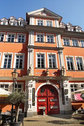 Barockes Gebäude mit roter Fassade, weißen Fensterrahmen und markantem Rundbogenportal mit Figurenschmuck