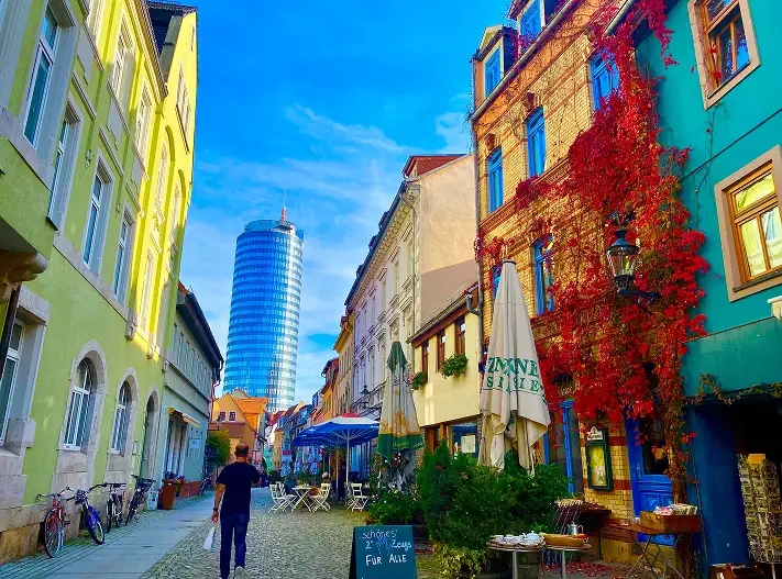 Eine bunte Altstadtgasse mit historischen Häusern, Cafés und Restaurants führt auf den modernen, verglasten JenTower im Hintergrund zu; ein Passant geht auf dem Kopfsteinpflaster durch die Szene.