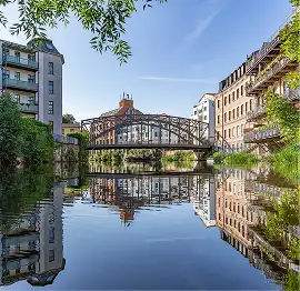 Alte Stahlbrücke über einem ruhigen Fluss, flankiert von historischen Wohnhäusern mit Spiegelung im Wasser.