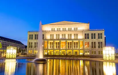 Beleuchtetes Opernhaus Leipzig am Abend mit Springbrunnen und Wasserspiegelung im Vordergrund.